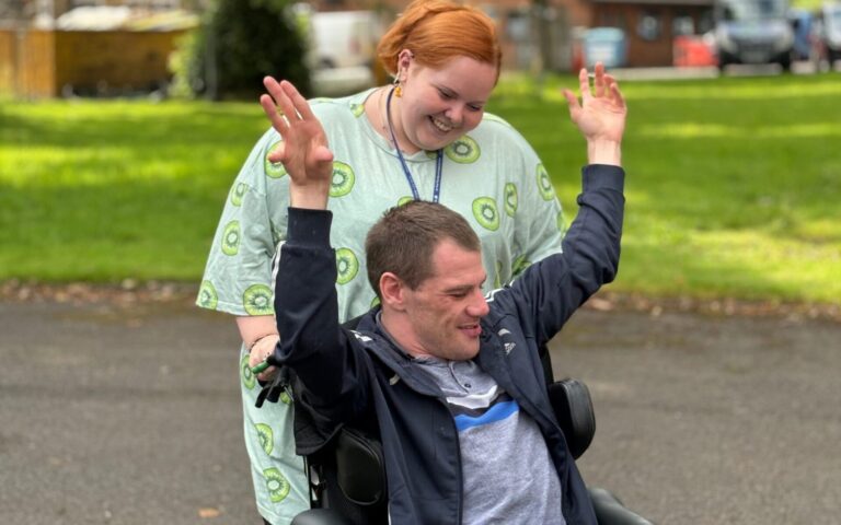Female support worker pushing man sat in his wheelchair with his arms up in the air. They are smiling together.