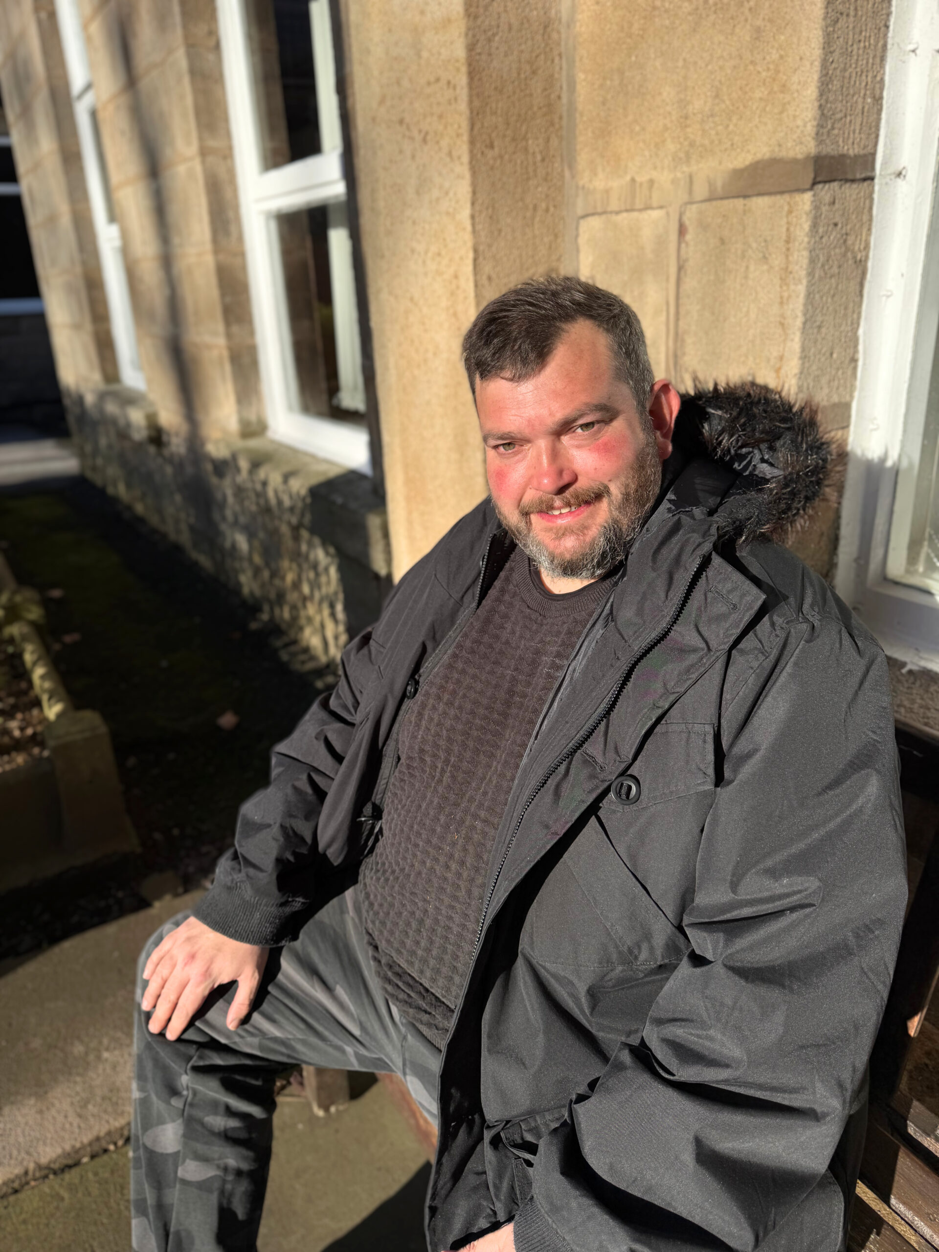 Photo of man smiling, he is dressed in dark clothing and sent on a wooden bench outside with the sun shining on him.