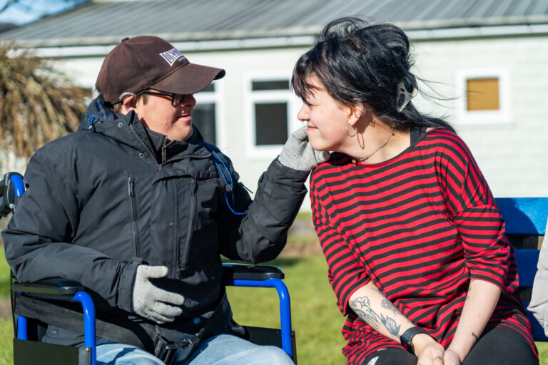 Man receiving learning disability support sits in a wheelchair outdoors, smiling and gently touching the face of a female support worker as they share a joyful moment.