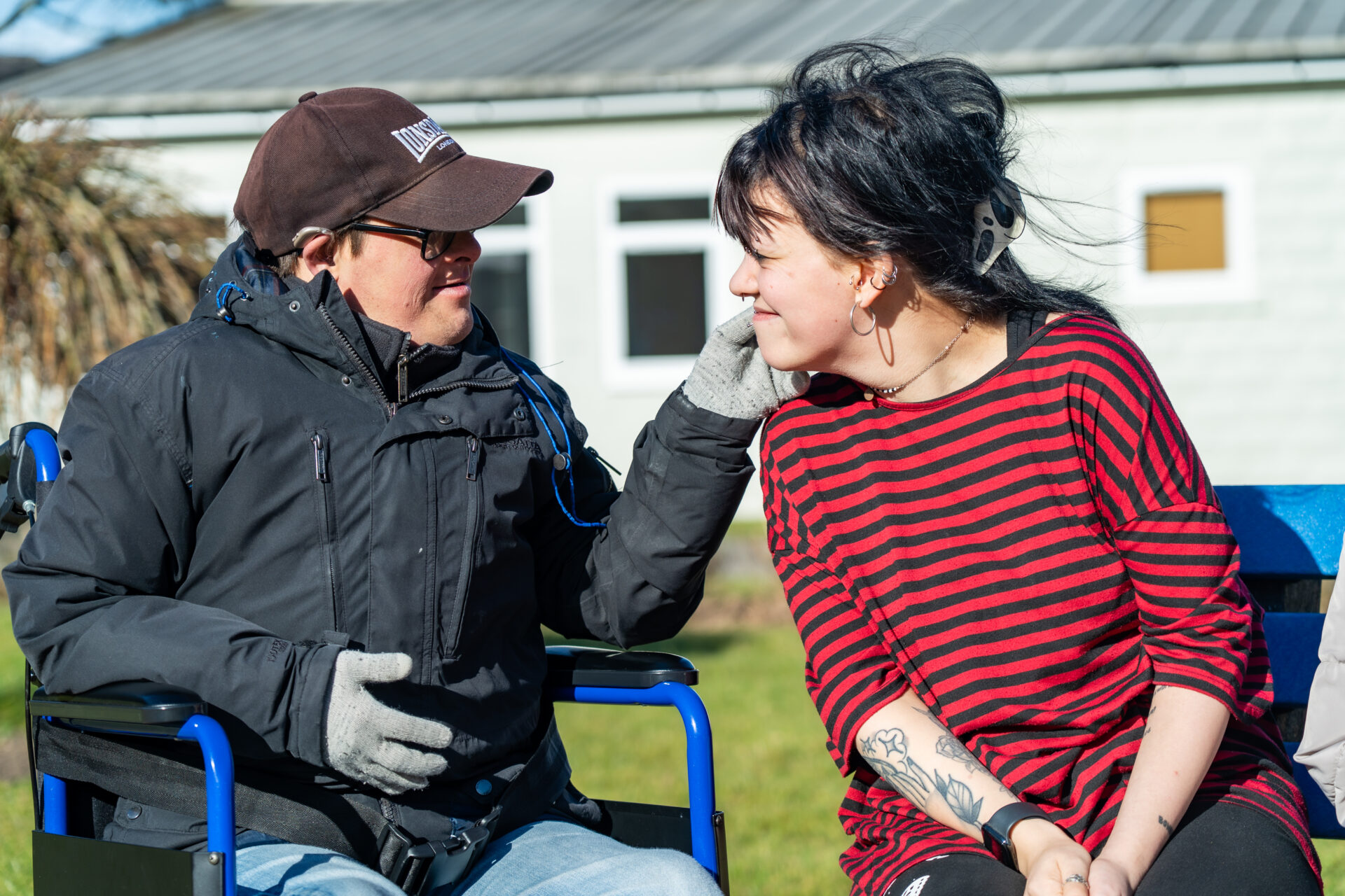 Man receiving learning disability support sits in a wheelchair outdoors, smiling and gently touching the face of a female support worker as they share a joyful moment.