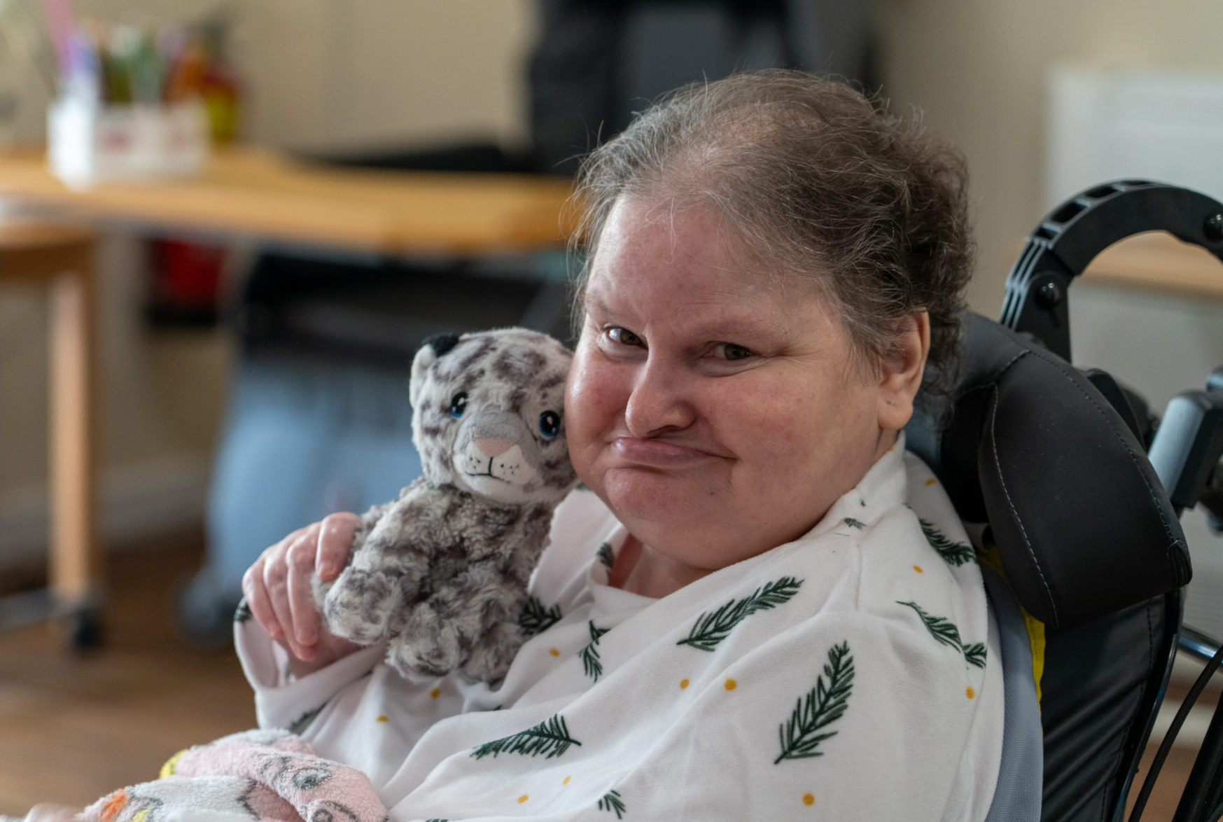 A senior woman in a wheelchair looking directly at the camera with a smile. She is holding a grey leopard soft toy close to her face.