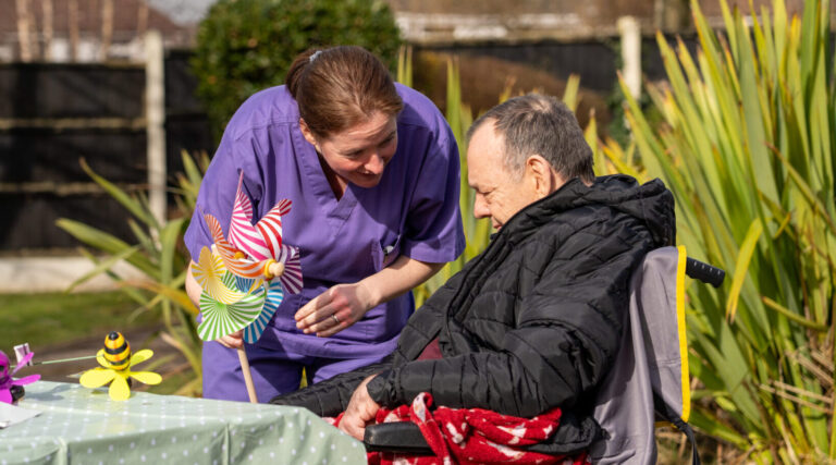 A nurse delivering nursing care, interacting with a man in a wheelchair pointing at a windmill spinner in the nursing home garden.